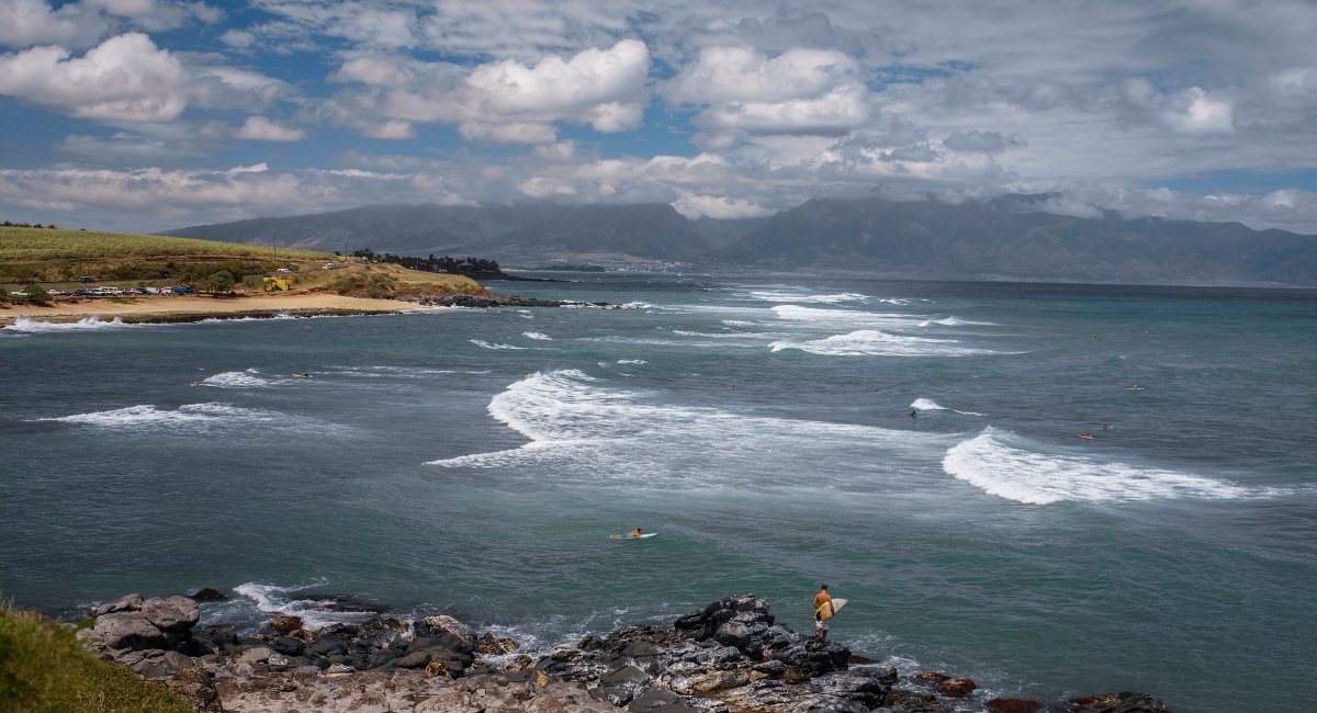 Scenic view of surfers riding waves on a vibrant day at Hoʻokipa Beach in Maui, Hawaiʻi, with rocky coastline in the foreground and dramatic mountain backdrop under a partly cloudy sky.