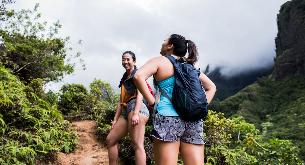 Two hikers smiling on a rainforest trail surrounded by ferns and dramatic mountain cliffs on Maui